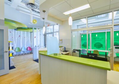 A bright, modern waiting room with a green reception desk, colorful chairs, and a children's play area.